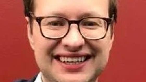 headshot of man wearing glasses, white button down and red and yellow patterned tie against a red background