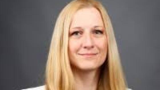 headshot of blonde woman wearing a light cream blazer and blue blouse against a dark gray background