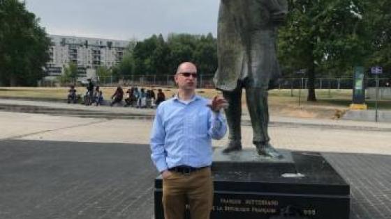 man in sunglasses, chinos and light blue button down standing in front of statue of man and a park in the background
