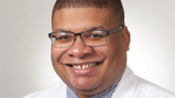 headshot of a man wearing glasses, red spotted tie, blue button down shirt, and a medical white coat