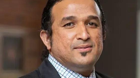headshot of man in dark gray suit with a dark blue spotted tie and blue and white plaid button down