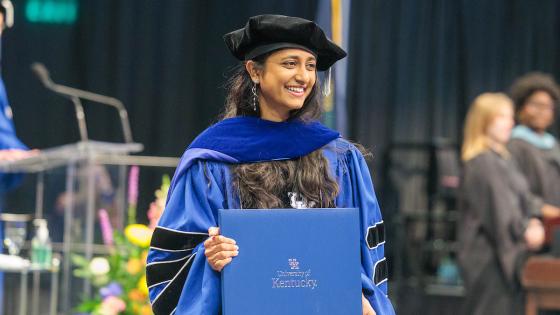 A UK doctoral student wearing blue graduation regalia holds their diploma and poses for a photo on the commencement stage.