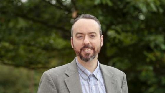 Aiyub Palmer posing for headshot wearing a light plaid button down shirt and a light brown jacket. 