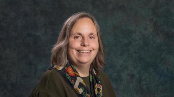 Karen Petrone wearing a dark caridgan and flowered scarf posed against a dark blue-green background.