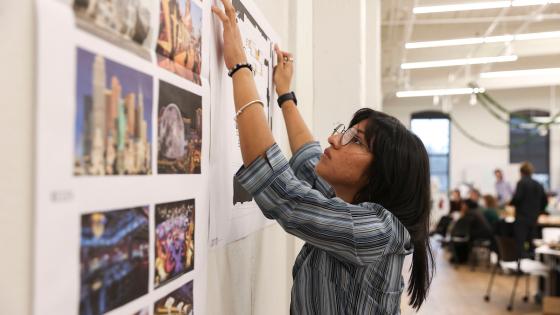 A student in a plaid shirt presses their hands against a poster to ensure it sticks to a wall, which is filled with other photos of designs and images.