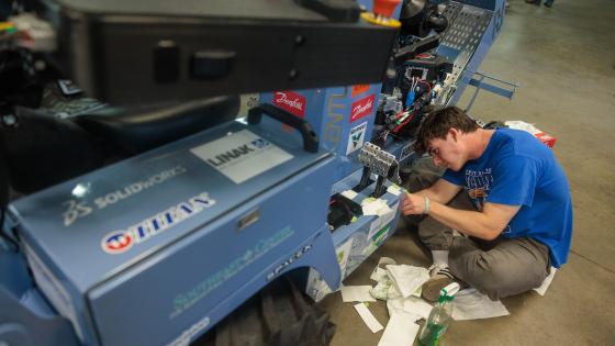 A student wearing a blue shirt sits on the ground next to a large tractor, affixing something to its side.
