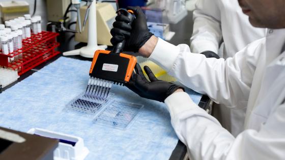 A person wearing a white lab coat uses a pipette to inject black liquid into a plastic container while at a lab bench filled with test tubes and equipment.