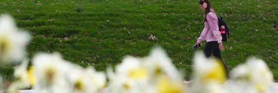 A student walking across the background with headphones and a pink sweater in springtime weather.