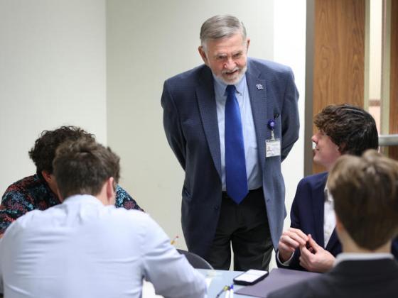 A man in a gray suit standing at a table talking to students.