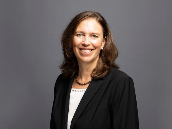 Woman with brown hair, black blazer and white shirt posing in front of a gray backdrop.