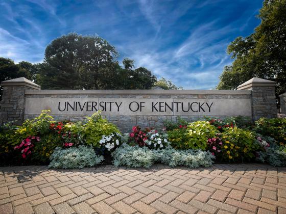 A large stone sign that says "University of Kentucky" with flower beds and a sidewalk in front and a blue sky in the background.