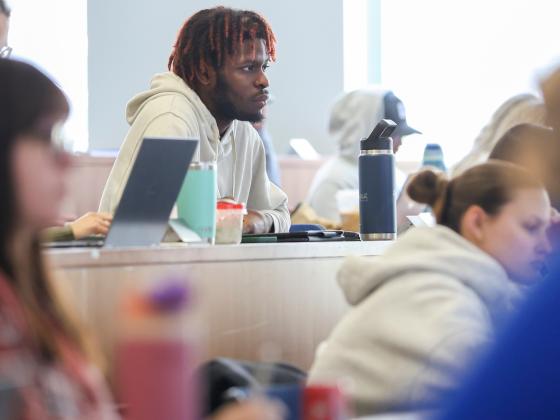 Student sitting in a classroom at a row of desks wearing a white hoodie.