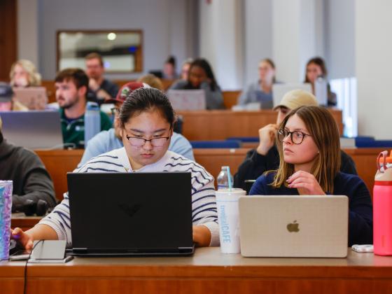 students sitting in class with laptops in tiered seating.