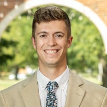 Logan Smart, wearing a tan suit and floral tie, posed in front of a brick arch and trees in the background.