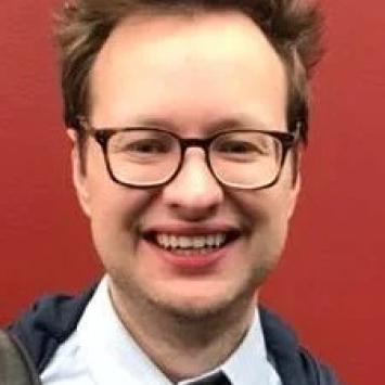 headshot of man wearing glasses, white button down and red and yellow patterned tie against a red background