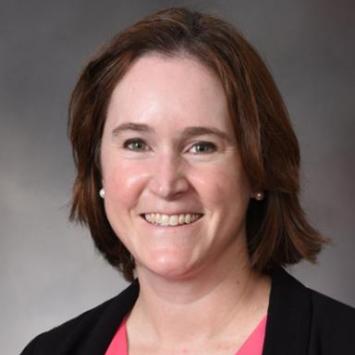 headshot of woman wearing coral shirt and a black jacket against gray background