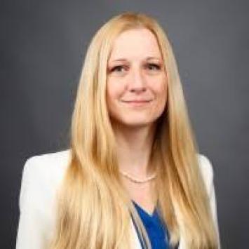 headshot of blonde woman wearing a light cream blazer and blue blouse against a dark gray background