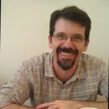 bearded man with glasses wearing a tan plaid shirt and arms crossed on top of a table against a beige wall