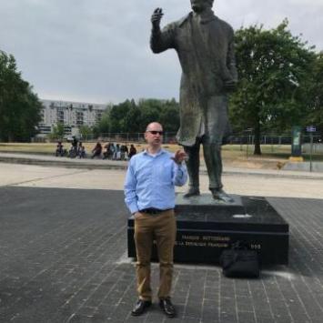 man in sunglasses, chinos and light blue button down standing in front of statue of man and a park in the background