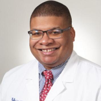 headshot of a man wearing glasses, red spotted tie, blue button down shirt, and a medical white coat