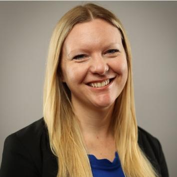 Blonde woman posing for headshot wearing a black blazer and UK blue shirt against a gray wall