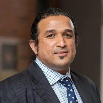 headshot of man in dark gray suit with a dark blue spotted tie and blue and white plaid button down