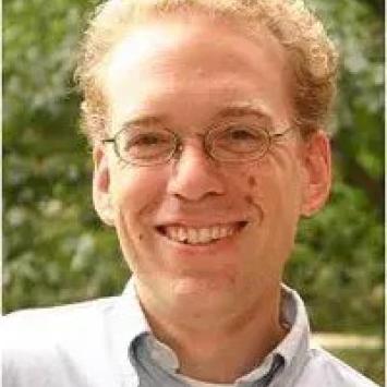 headshot of man in glasses and light blue button down against greenery and leaves