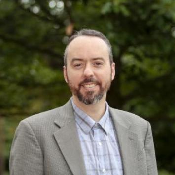 Aiyub Palmer posing for headshot wearing a light plaid button down shirt and a light brown jacket. 