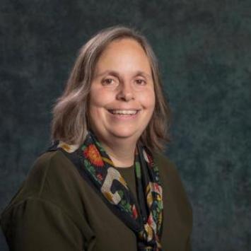 Karen Petrone wearing a dark caridgan and flowered scarf posed against a dark blue-green background.