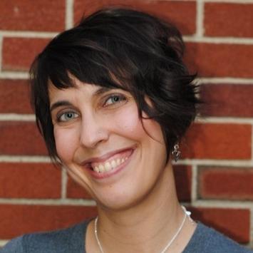 Woman with short brown hair smiling and posing against a brick wall