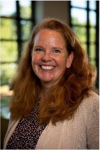 headshot of woman with red hair smiling wearing a printed purple blouse with a neutral sweater overtop. 