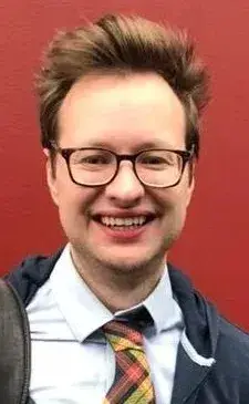 headshot of man wearing glasses, white button down and red and yellow patterned tie against a red background