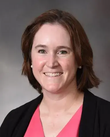 headshot of woman wearing coral shirt and a black jacket against gray background