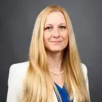headshot of blonde woman wearing a light cream blazer and blue blouse against a dark gray background