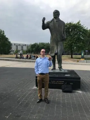 man in sunglasses, chinos and light blue button down standing in front of statue of man and a park in the background