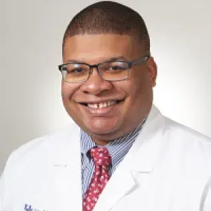 headshot of a man wearing glasses, red spotted tie, blue button down shirt, and a medical white coat