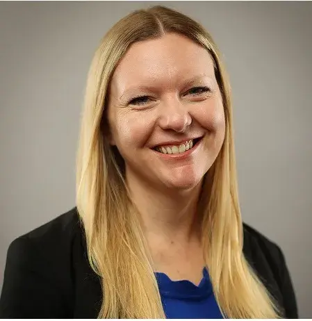 Blonde woman posing for headshot wearing a black blazer and UK blue shirt against a gray wall