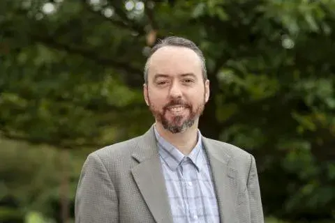 Aiyub Palmer posing for headshot wearing a light plaid button down shirt and a light brown jacket. 