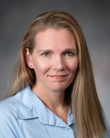 Jill Clemons posing for a headshot wearing a light blue collared shirt against a gray background. 
