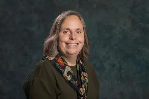 Karen Petrone wearing a dark caridgan and flowered scarf posed against a dark blue-green background.