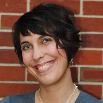 Woman with short brown hair smiling and posing against a brick wall